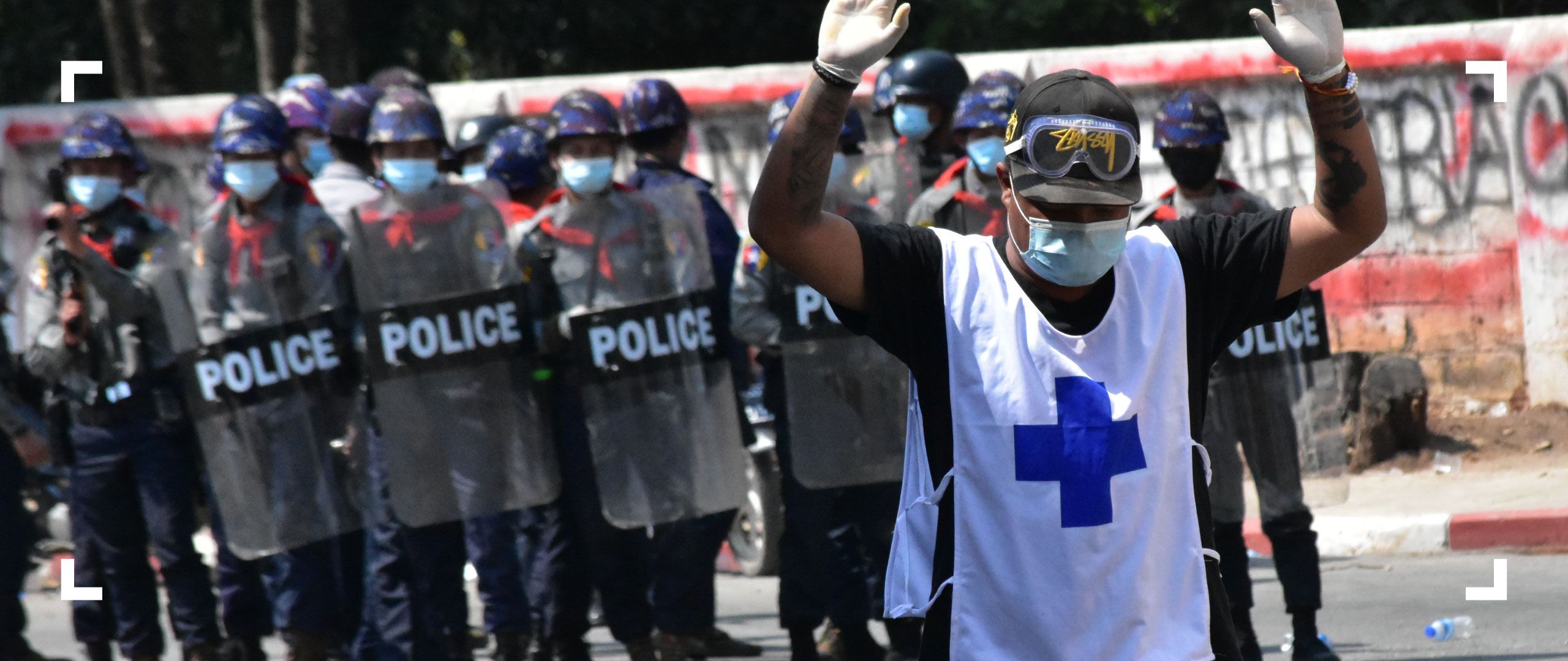 Photograph of a man with his hands over his head, and his back turned to a group of militarized police officers.